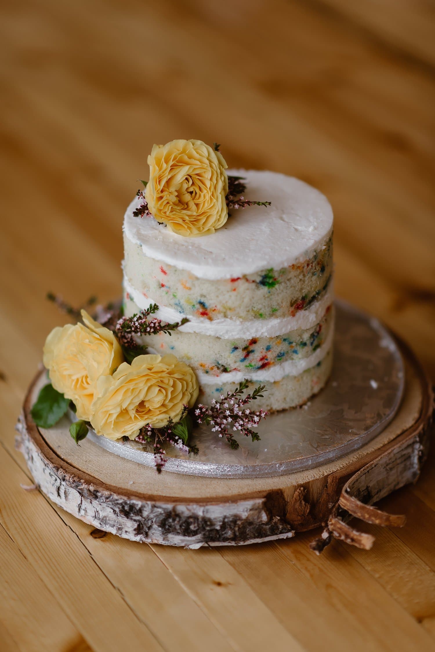 Confetti cake, with white frosting, on a tree stump platter. There are yellow roses next to the cake. 