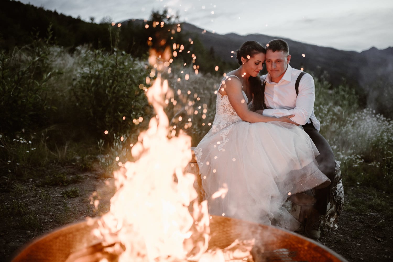 Bride and groom sit together during blue hours in Alaska, embracing, in front of a bonfire.