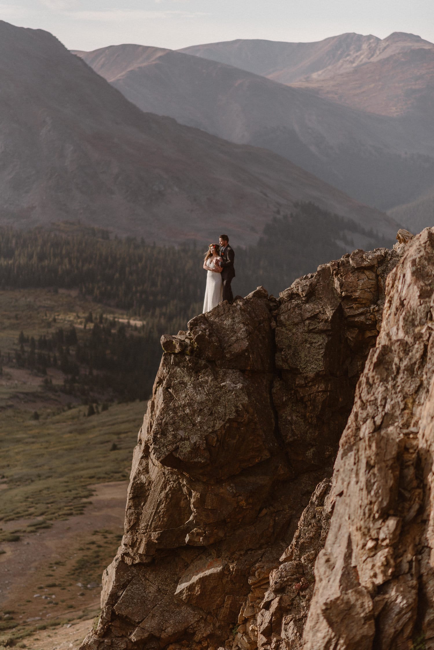 Bride stands in front of groom on cliff as they hold hands and look out at the view. 