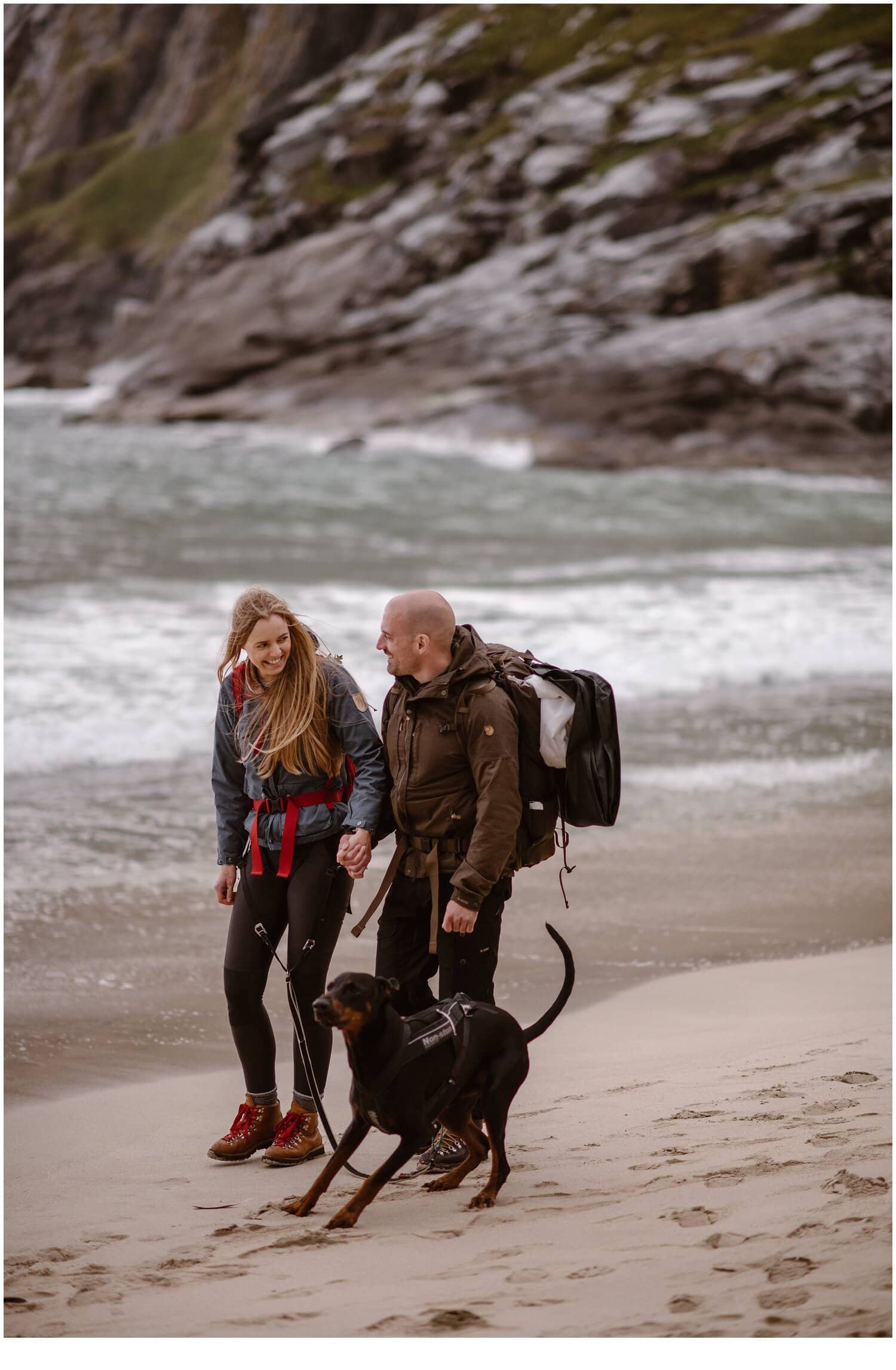 Bride and groom hold hands and walk along a beach with their dog in Norway. 
