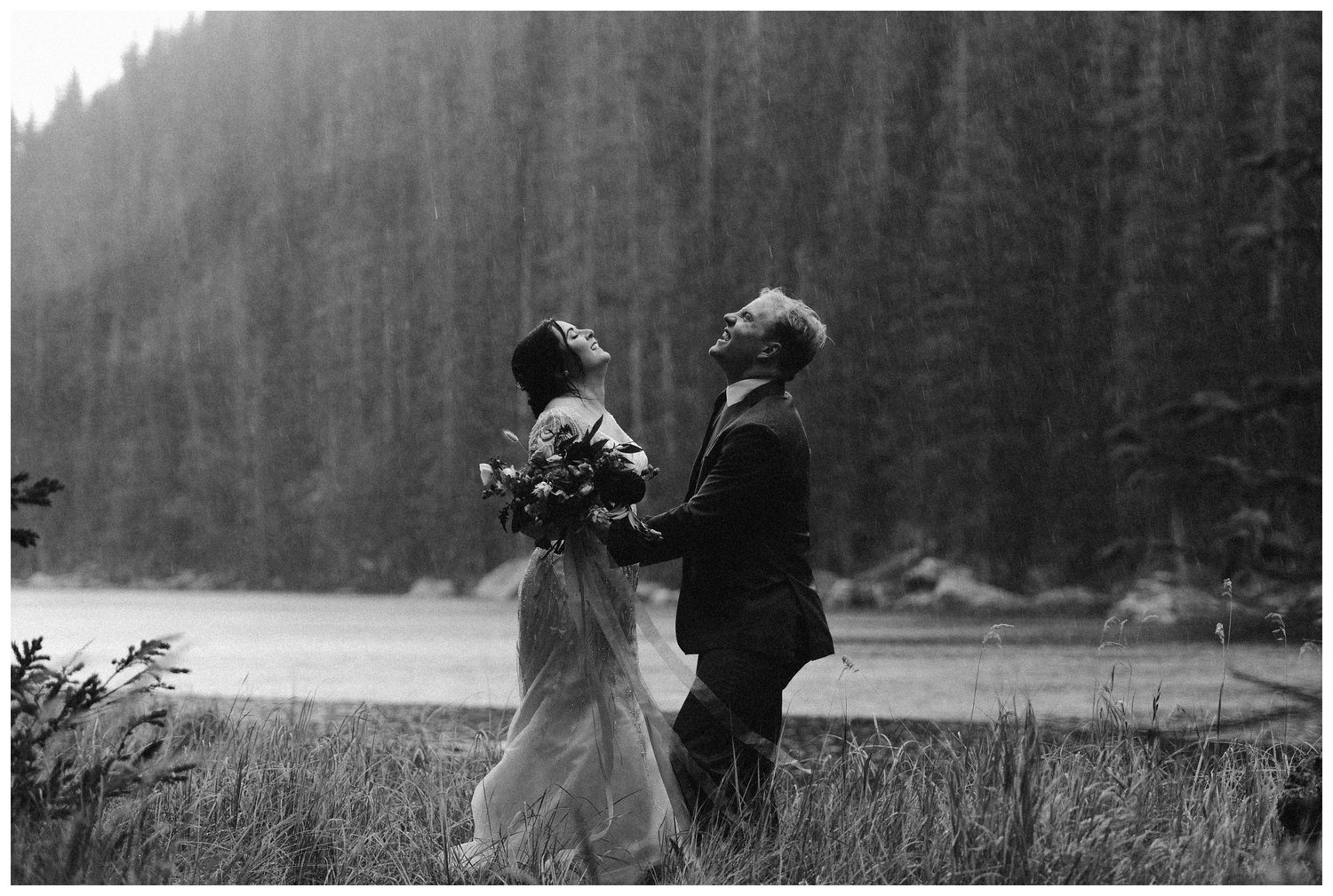 Bride and groom look up as it rains on them with lake and forest behind them. 
