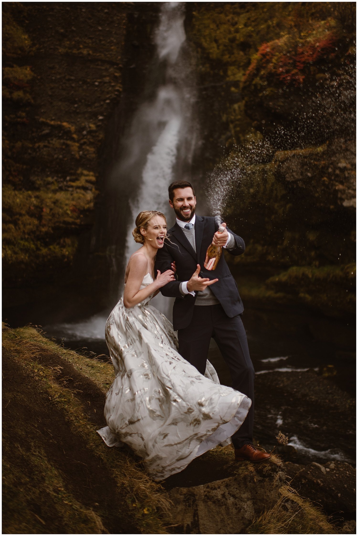 Bride and groom pop champagne in front on waterfall. 