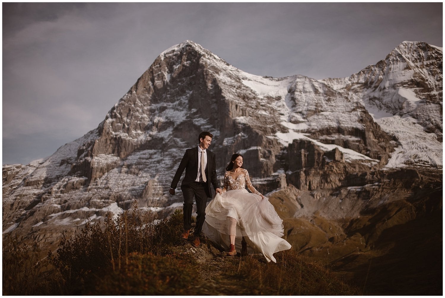 Bride and groom walk along trail with snow-capped mountains in background. 