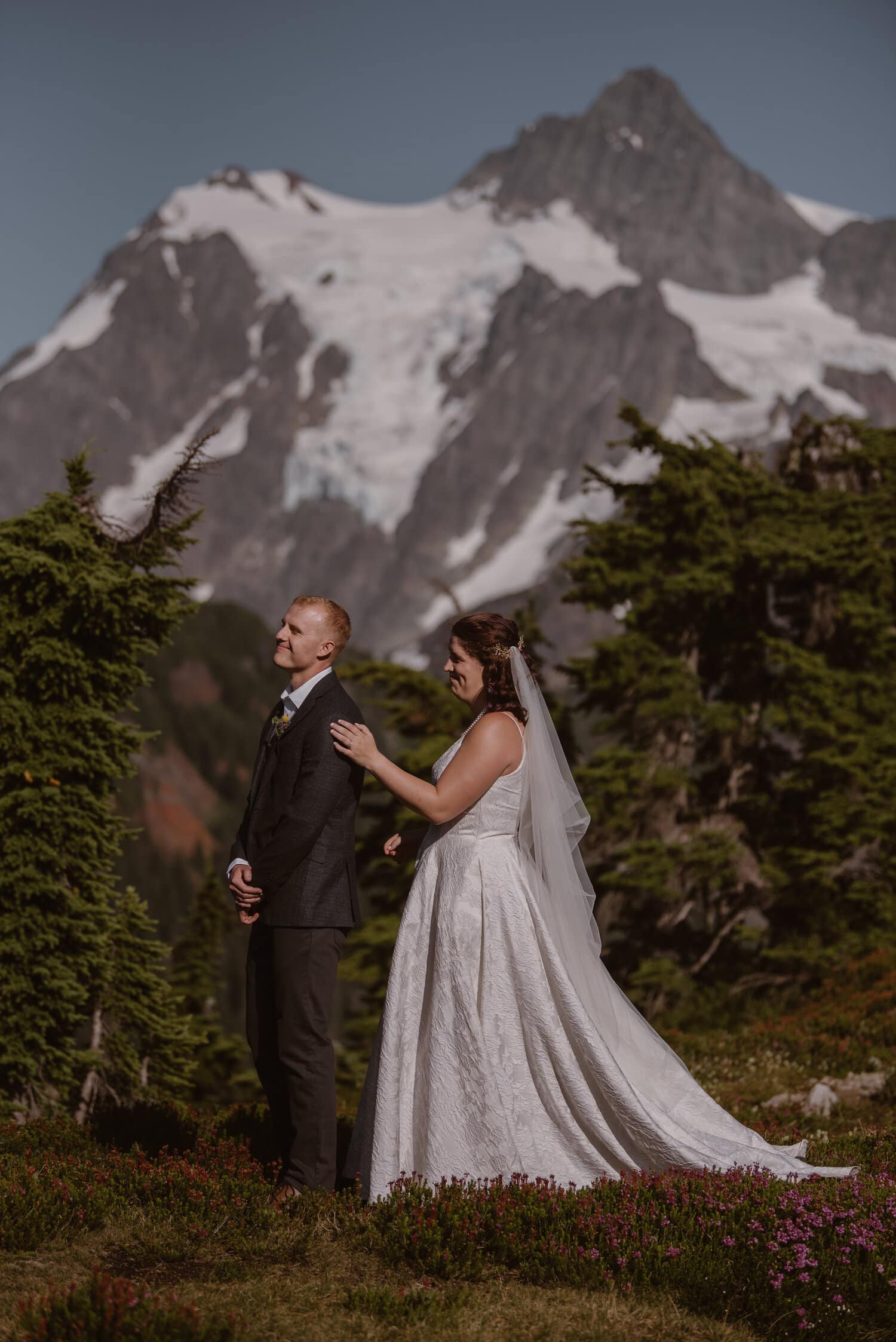 Bride approaches groom for a first look on their elopement day in Washington. Mount Shuksan is in the background, and there are trees and wildflowers around them. 