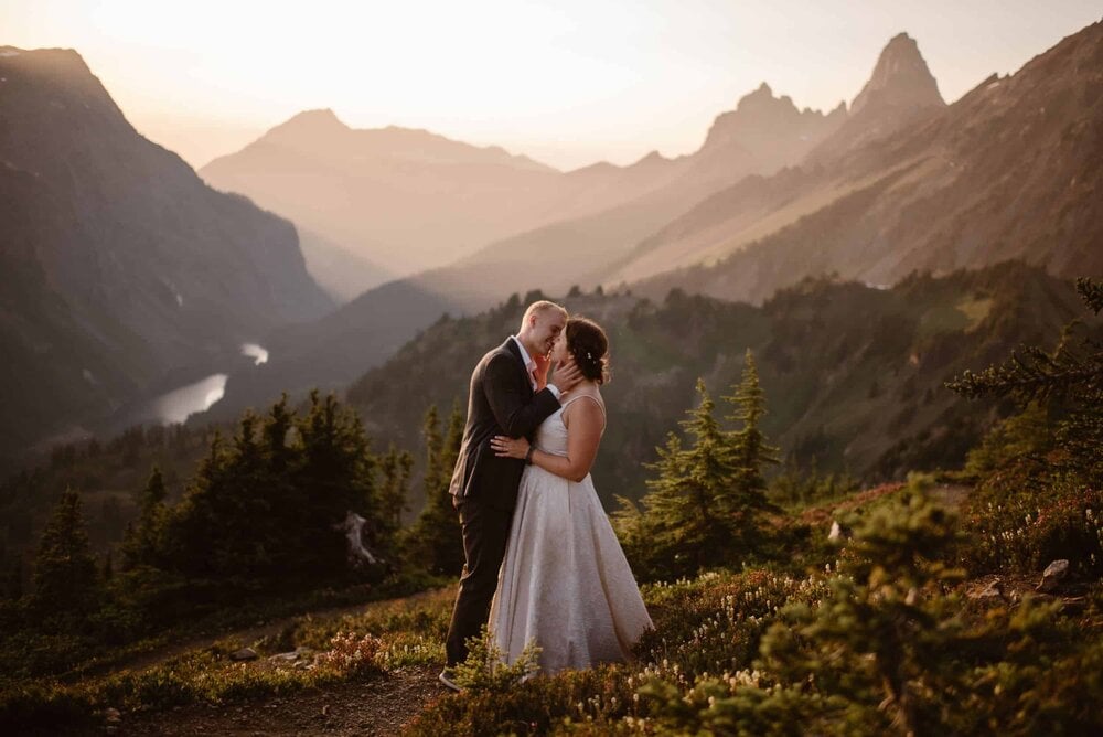 Bride and groom kiss on a grassy hill with wildflowers, in Washington at sunset. There are mountains in the background. 