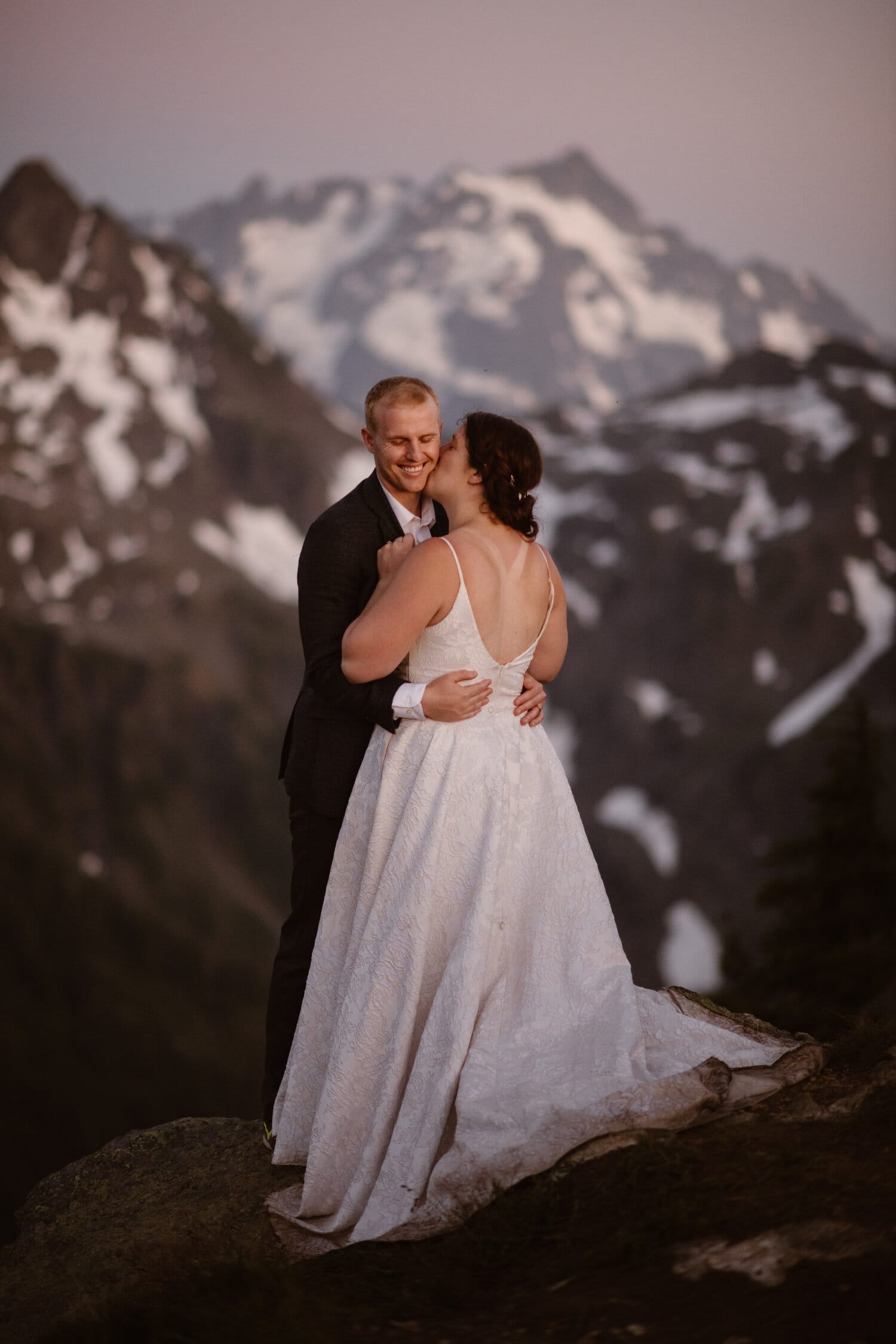 Bride kisses groom on the cheek on their elopement day in Washington. There are snow-capped mountains in the background. 