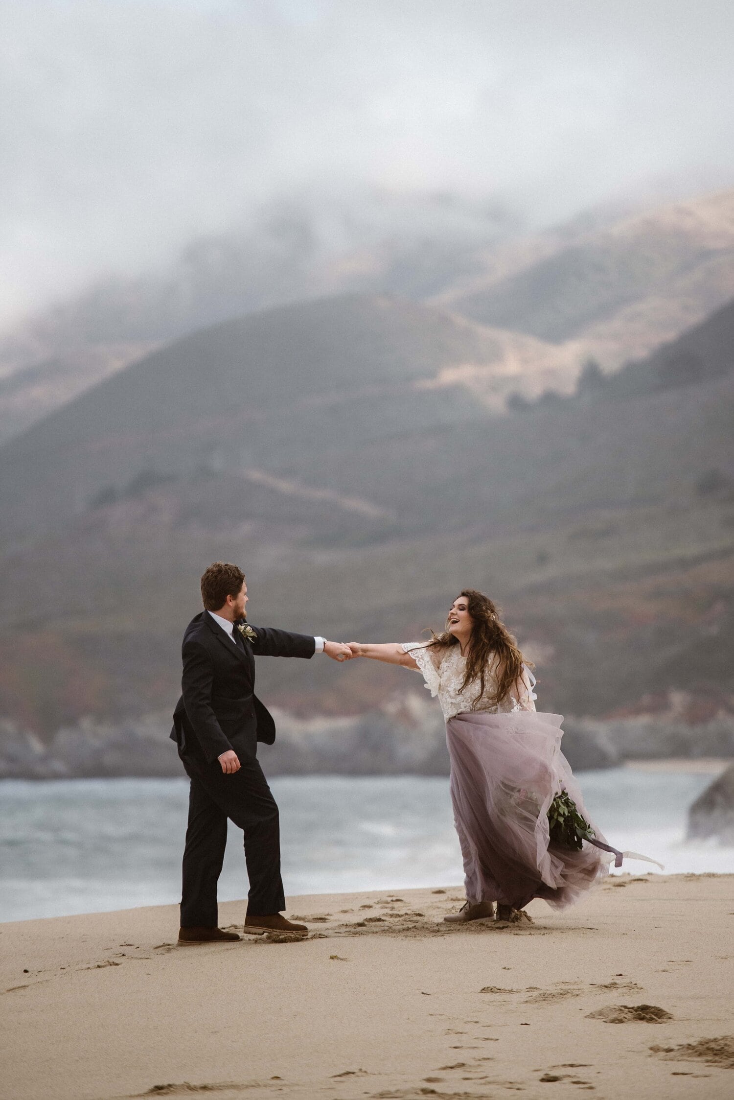 Bride and groom hold hands while smiling on their elopement day. They are standing on the beach in Big Sur, California. 