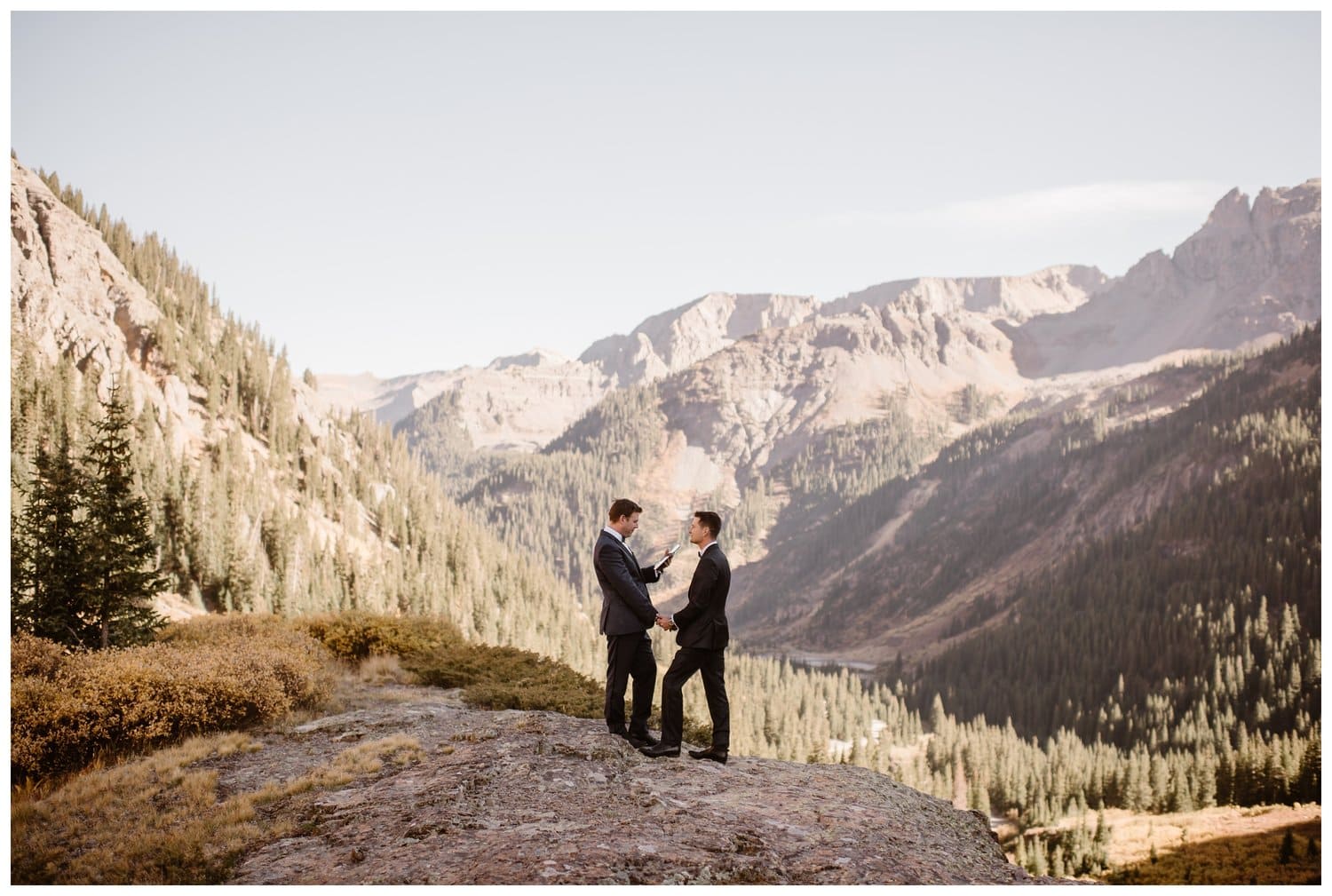 two grooms read their vows to each other with forest and mountains in the background. 