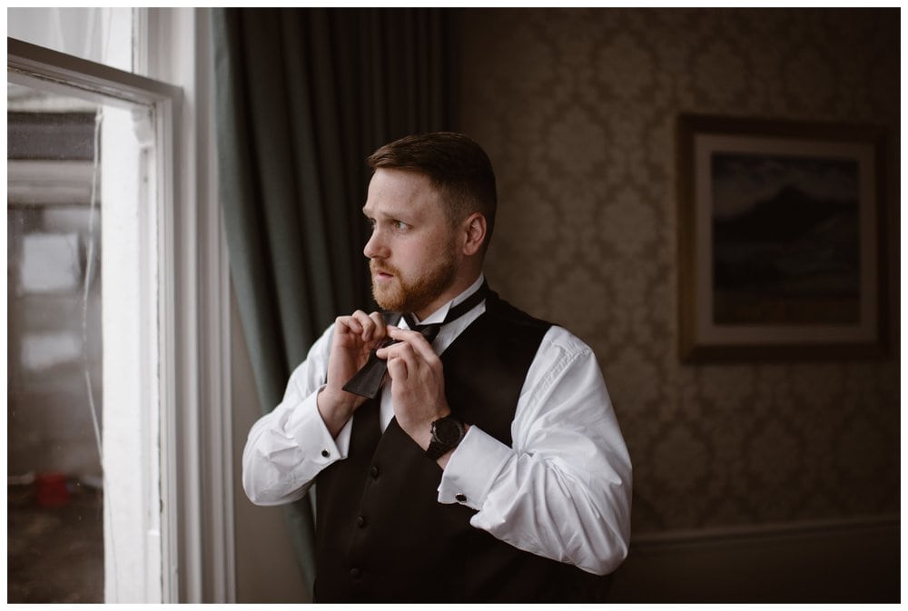 Groom ties his bow tie in a hotel while looking out a window. 