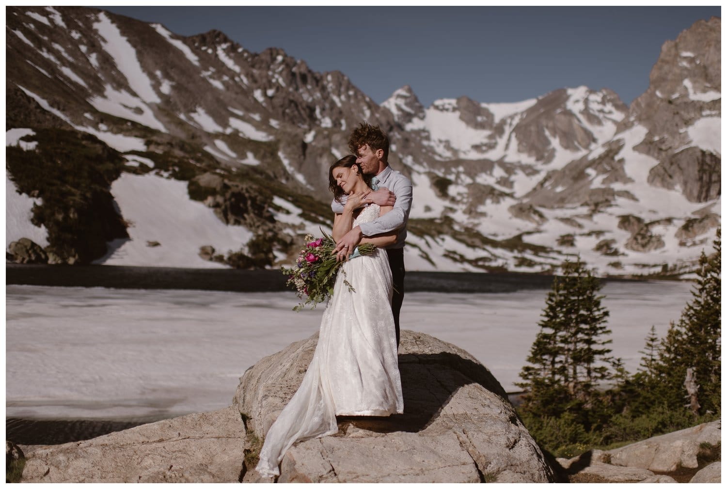 Bride and groom embrace while standing on a rock, in front of an alpine lake, at the Indian Peaks in Colorado. There are trees and snow-capped mountains in the background. 
