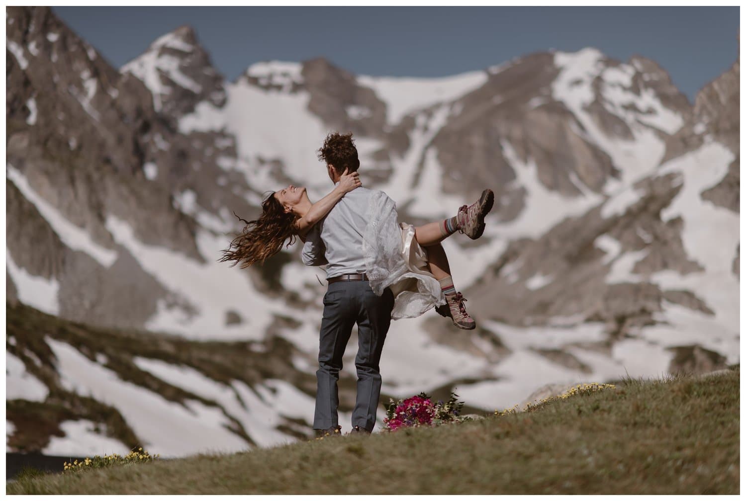 Groom holds bride on a ridge with wildflower, at the Indian Peaks in Colorado. There are snow-capped mountains in the background. 