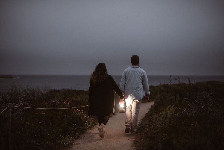 Bride and groom are walking on a trail next to the ocean and the bride is holding a lantern, in Monterey, California.