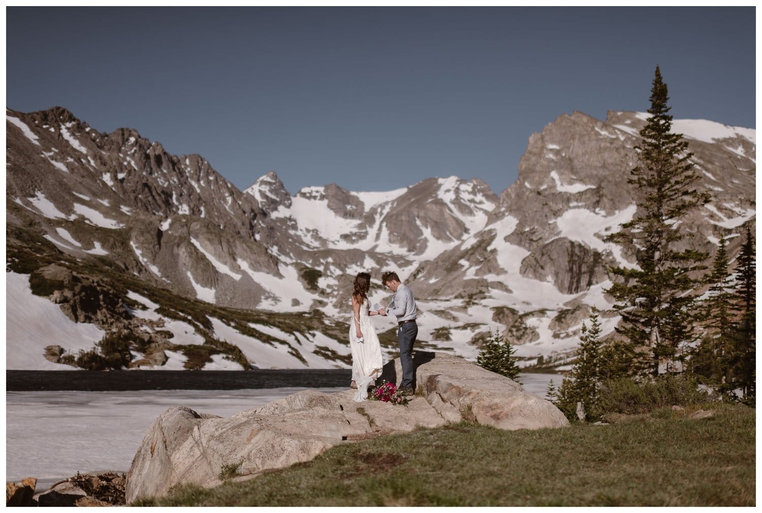 Bride and groom stand in front of an alpine lake, at the Indian Peaks in Colorado, during their intimate elopement ceremony. 