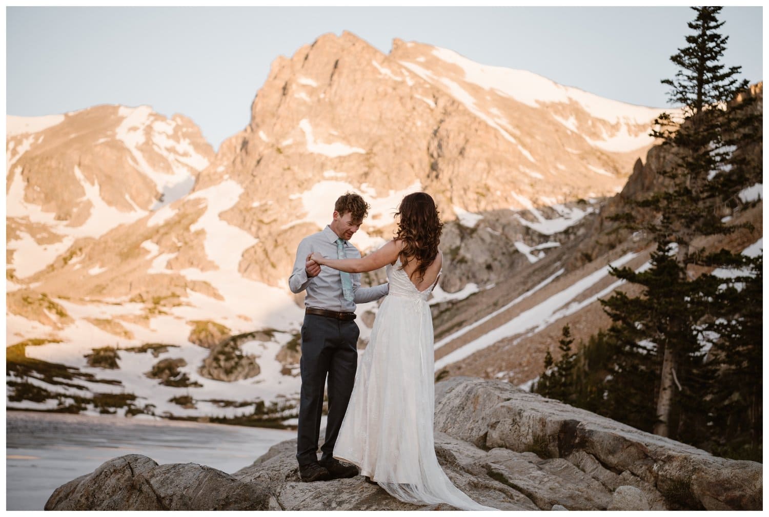Bride and groom share a first look on their elopement day at the Indian Peaks in Colorado. There is an alpine lake and snow-capped mountains with alpenglow in the background.