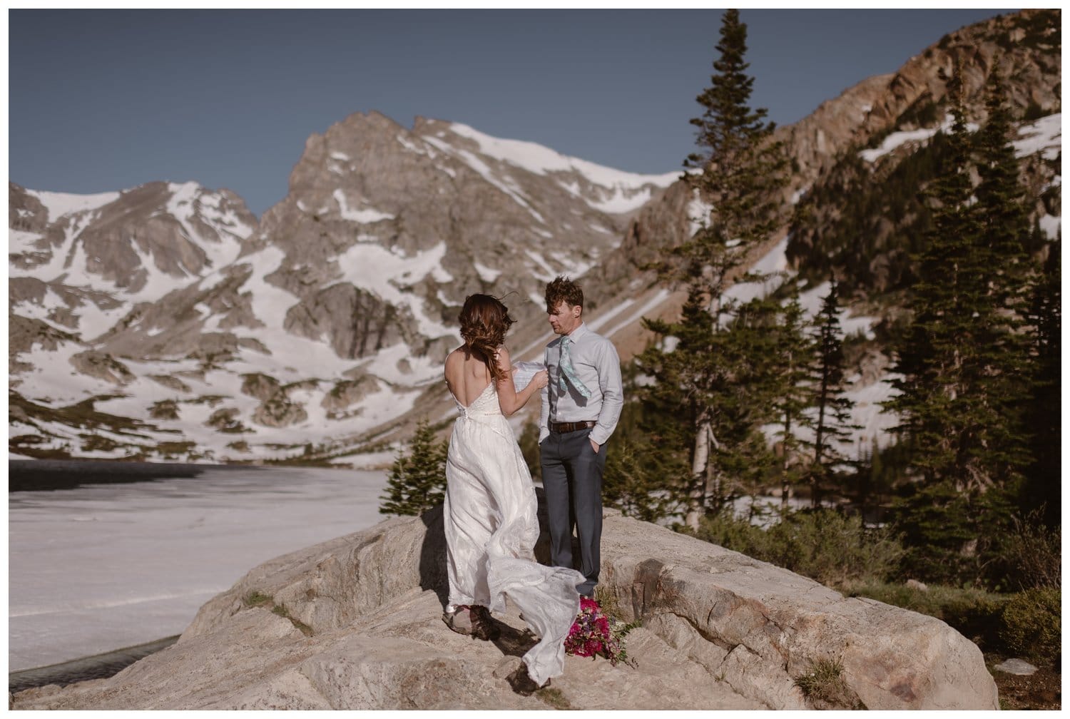 Bride reads her vows in front of an alpine lake, during intimate elopement ceremony at the Indian Peaks in Colorado. There are trees and snow-capped mountains in the background. The bride's dress is flowing in the wind. 