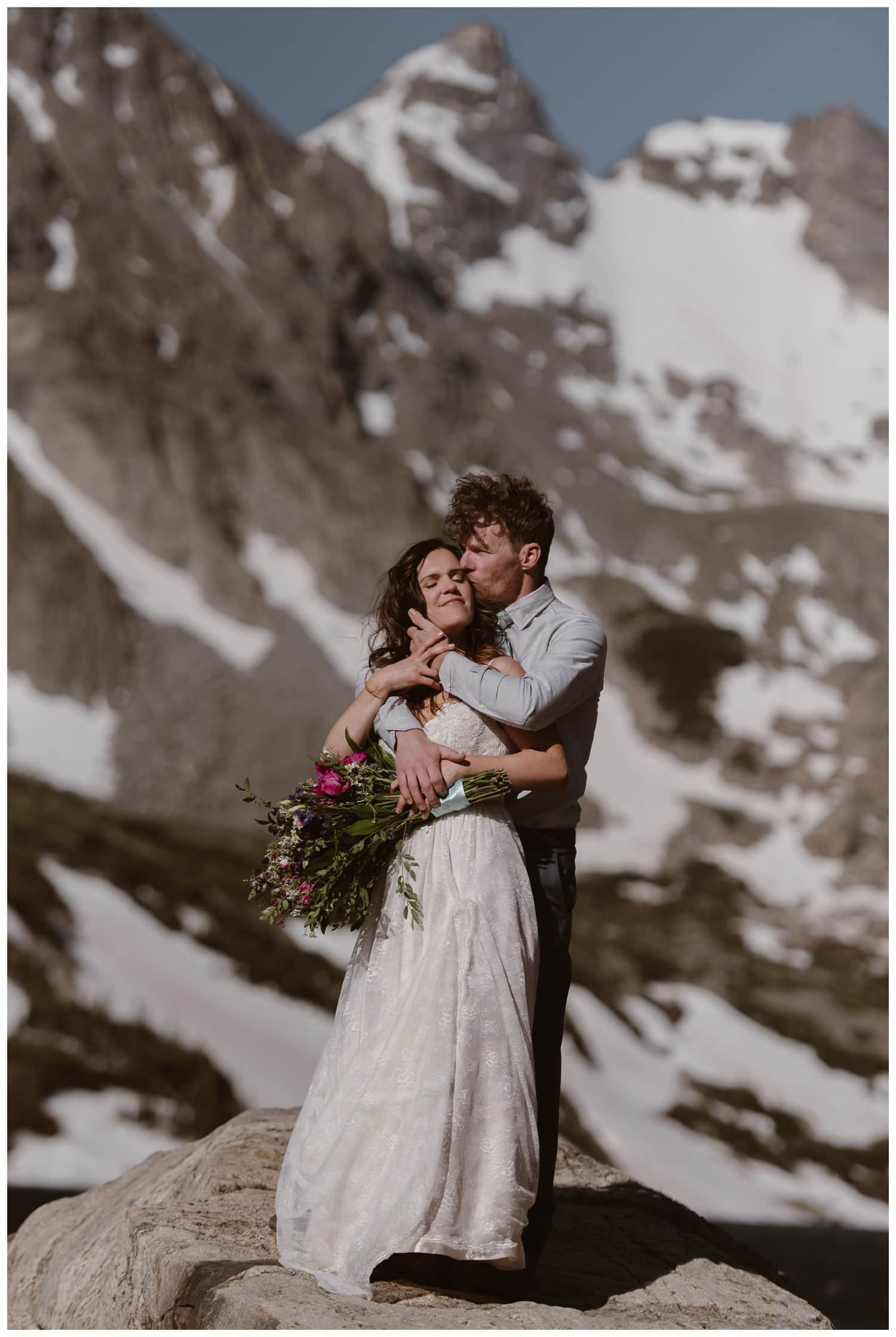 Bride and groom embrace, and groom kisses bride on the cheek at the Indian Peaks in Colorado. 