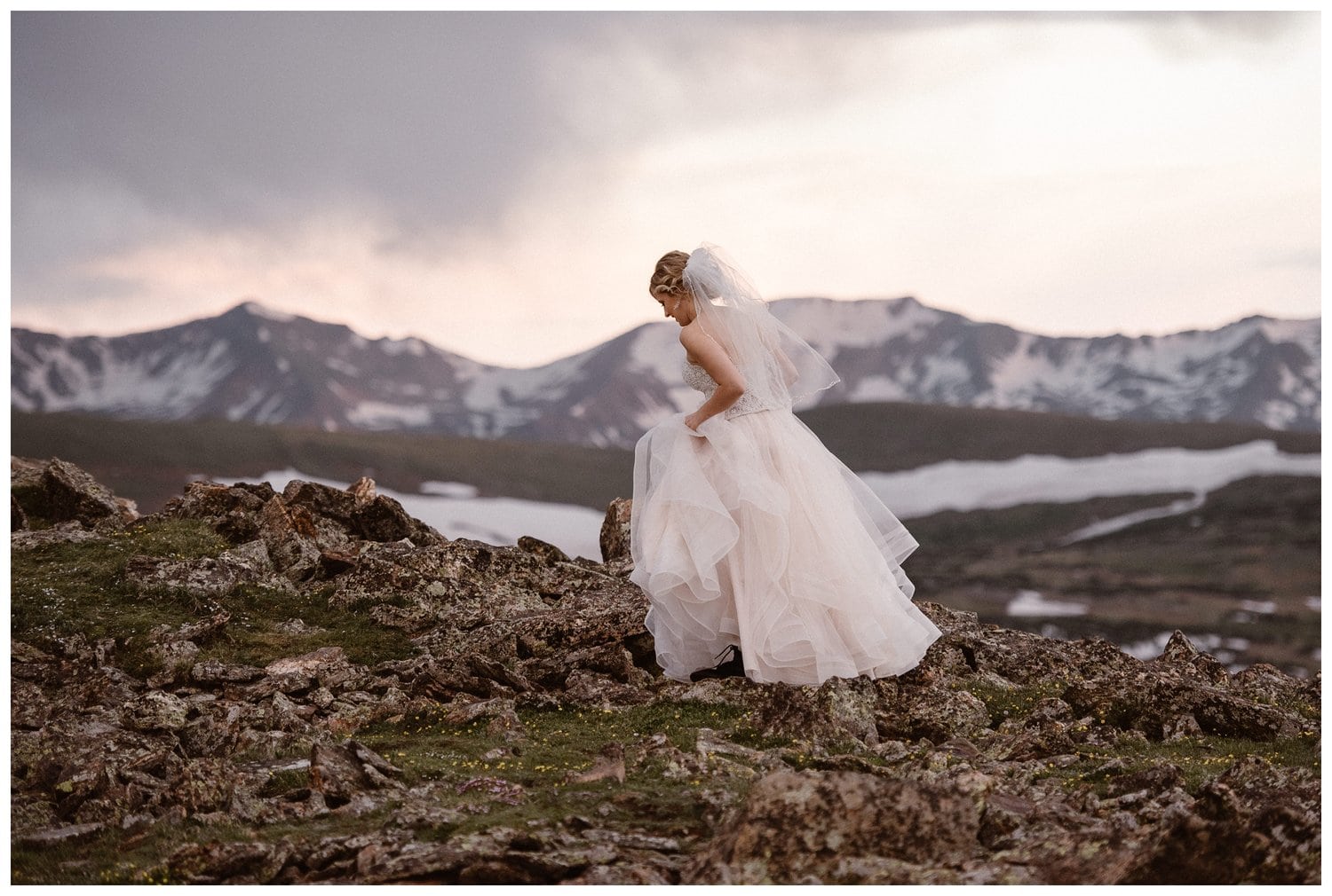 Bride wearing two-piece white dress and veil at Trail Ridge Road in Rocky Mountain National Park, Colorado. There are snow-capped mountains in the background. 
