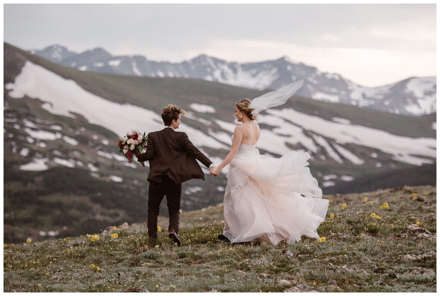 Two brides hold hands and walk through meadow with yellow wildflowers at Trail Ridge Road in Rocky Mountain National Park Colorado. There are snow-capped mountains in the background. 