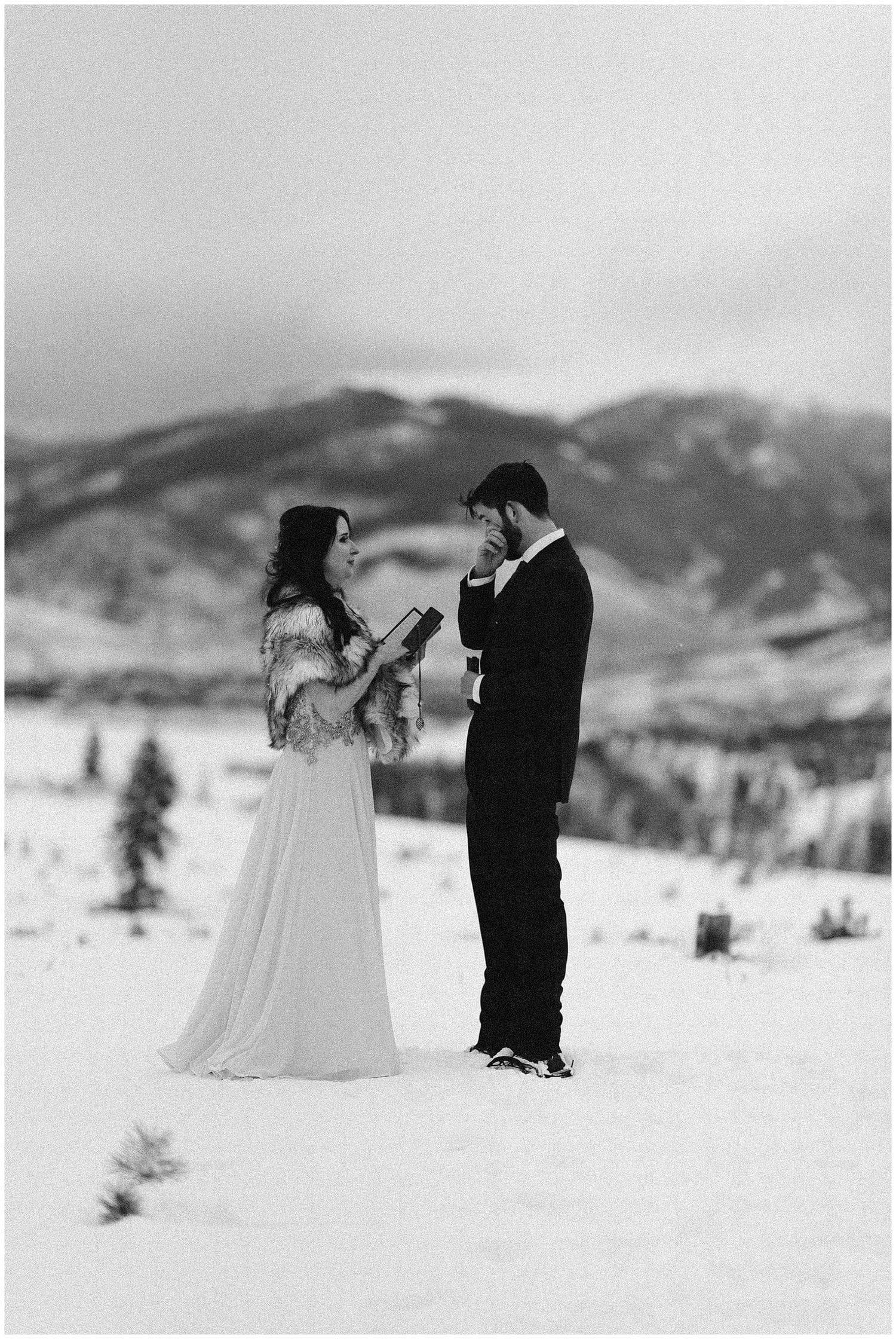 Bride and groom read their vows during intimate elopement ceremony. They are surrounded by snow, and there are mountains in the background at Sapphire Point, Colorado. 