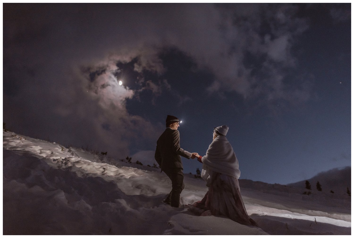 Bride and groom walk up a snow-covered hill, with headlamps at night. 
