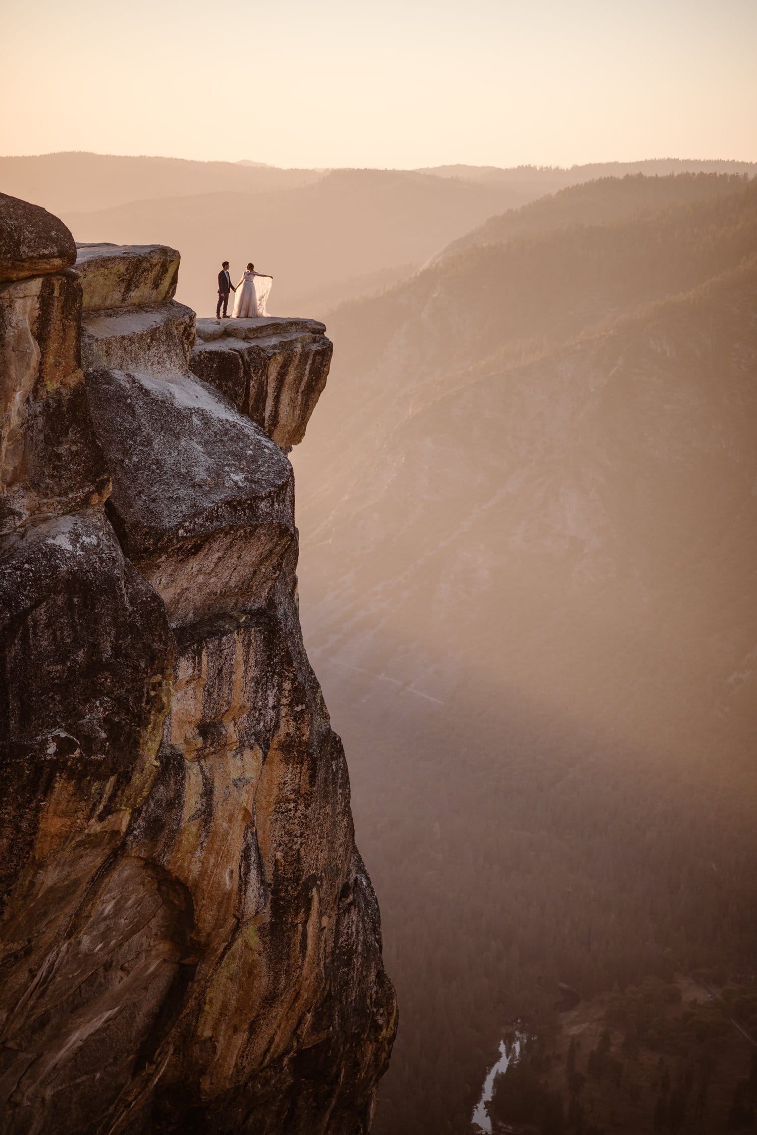 Bride and groom stand on cliff, holding hands, with mountains in background. 
