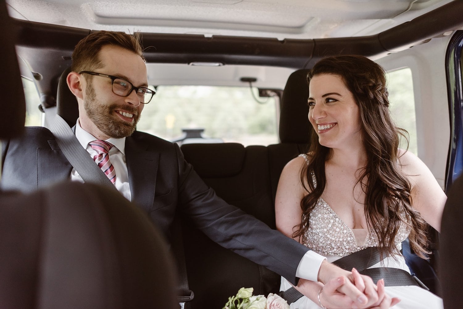 Bride and groom hold hands while sitting in Jeep. 
