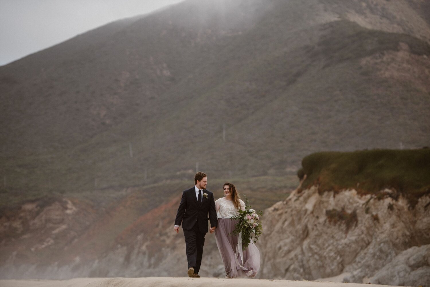 Bride and groom hold hands and walk along the beach on their elopement day in Big Sur, California.