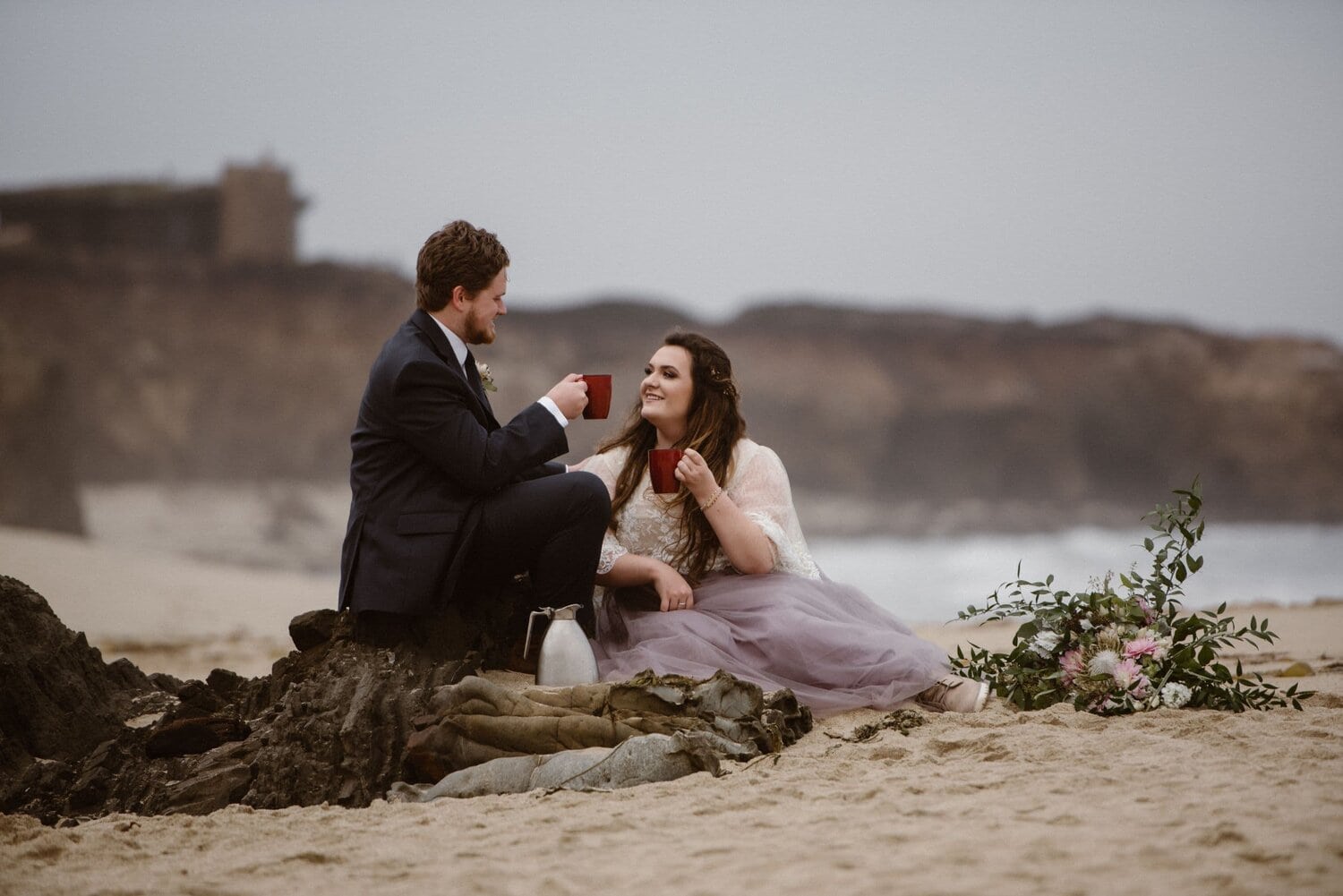 Bride and groom sitting on the beach at sunrise and drink coffee, in Big Sur, California.