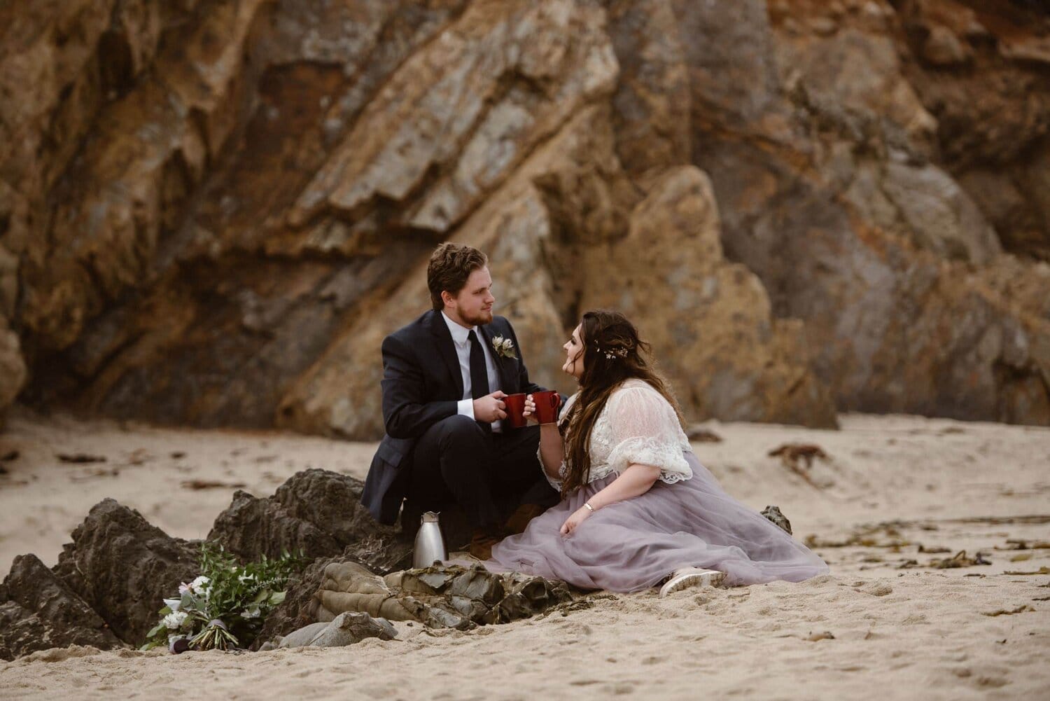 Bride and groom sitting on the beach at sunrise and drink coffee, in Big Sur, California.