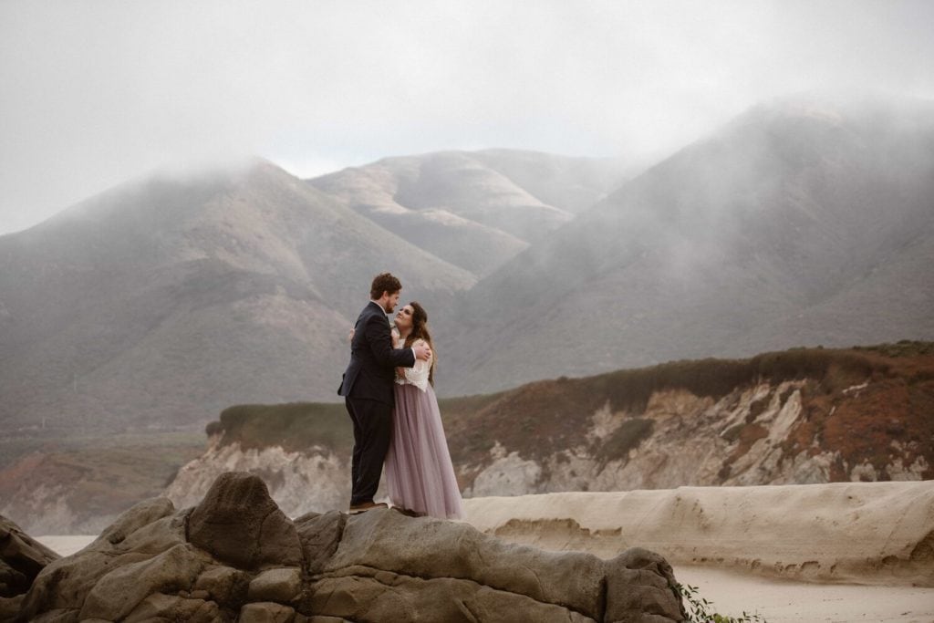 Bride and groom embrace while standing on rocks, next to the beach, in Big Sur, California. The sky is cloudy and there are mountains in the background. 