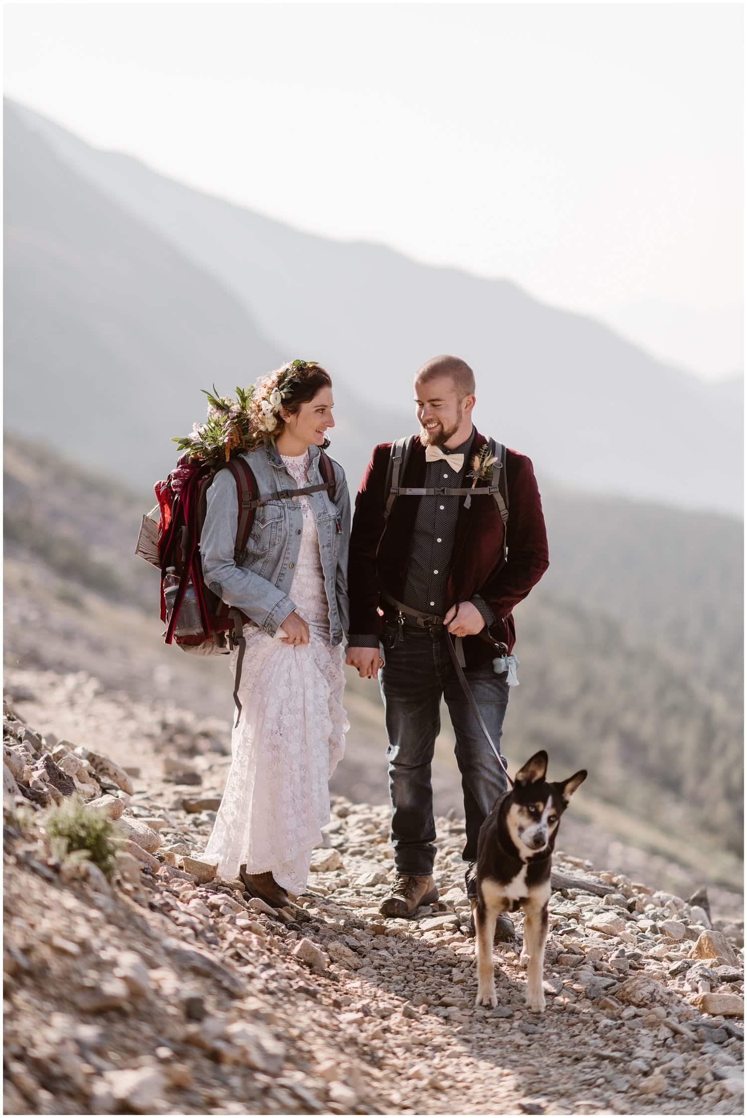 A bride and groom walk hand in hand while hiking with their dog on their wedding day.