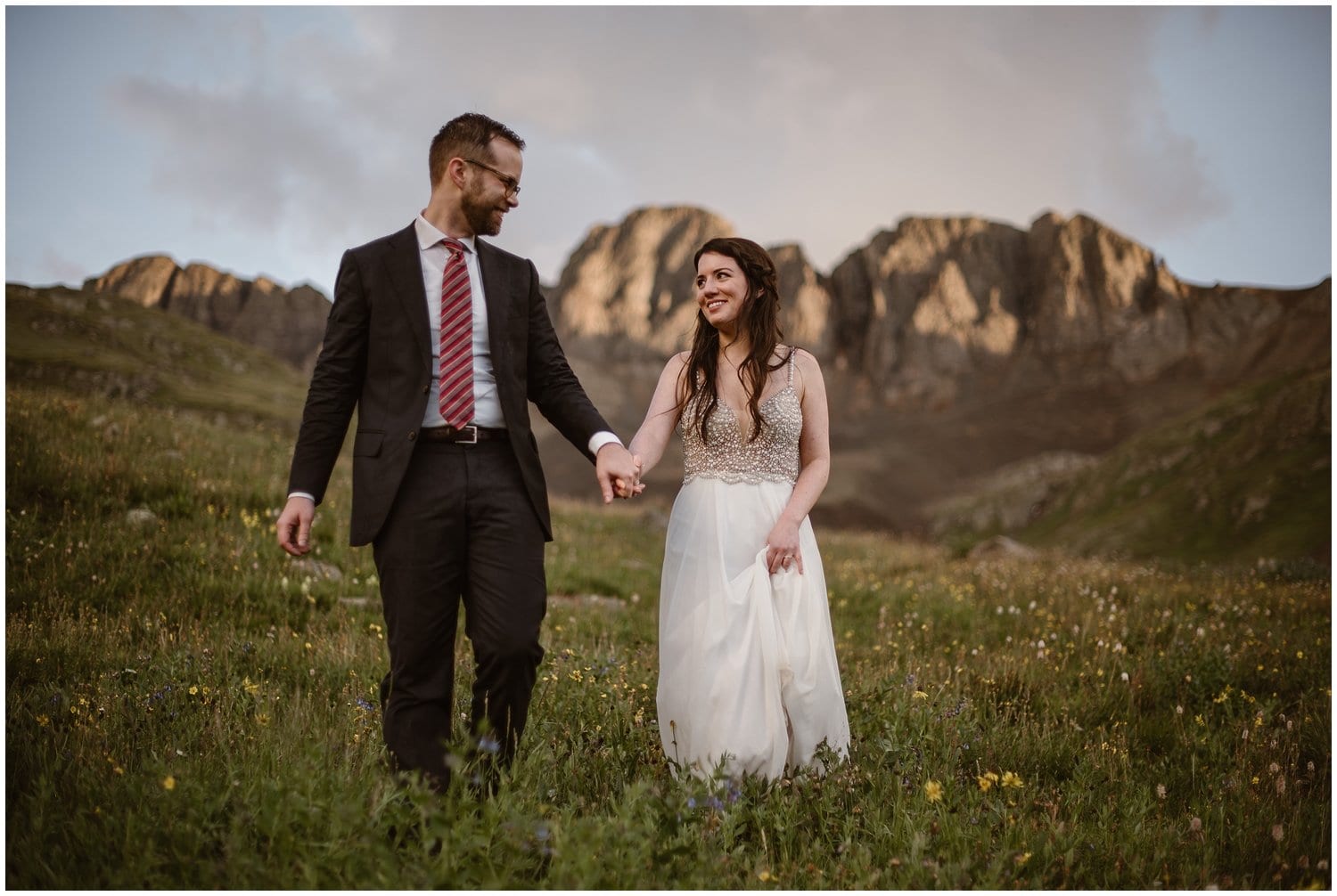 Bride and groom hold hands and walk through a field of wildflowers in the San Juan Mountains. 
