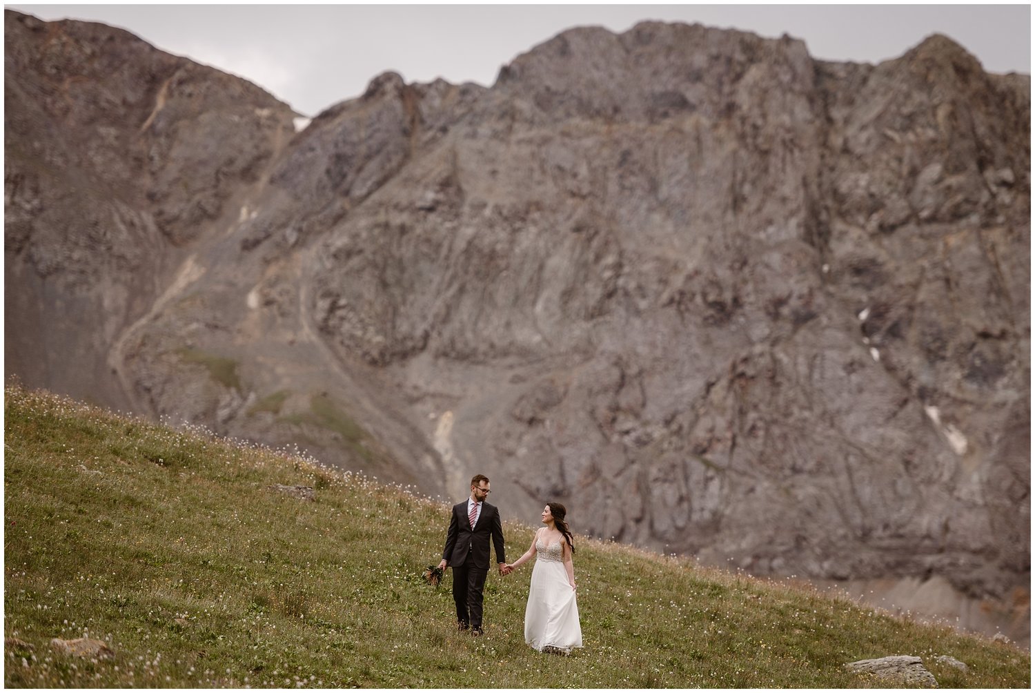 Bride and groom walk through field, with San Juan Mountains in the background. 