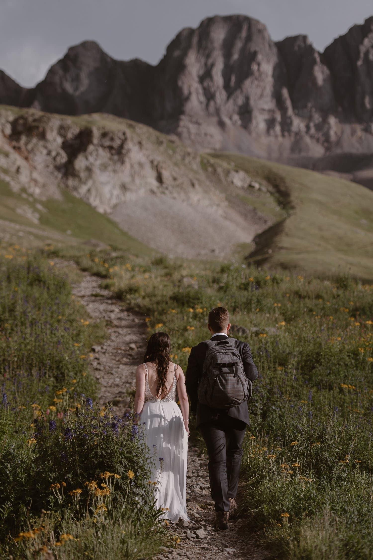 Bride and groom walk on trail, surrounded by wildflowers. 