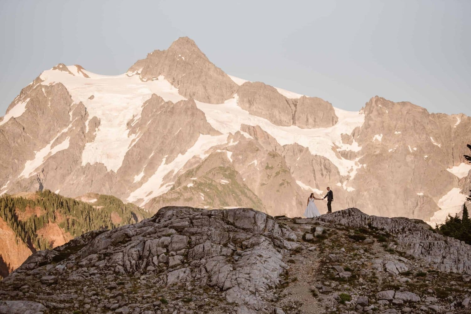 Bride and groom hold hands and are standing on a mountain ridge at the base of Mount Shuksan in Washington. 