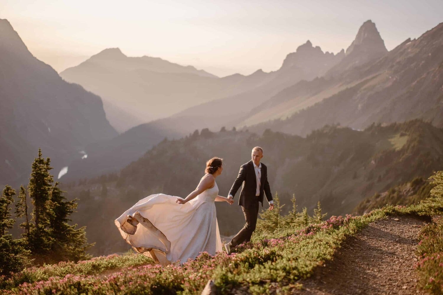 Bride and groom hold hands and walk on a grassy hill with wildflowers in Washington at sunset. There are mountains in the background. 
