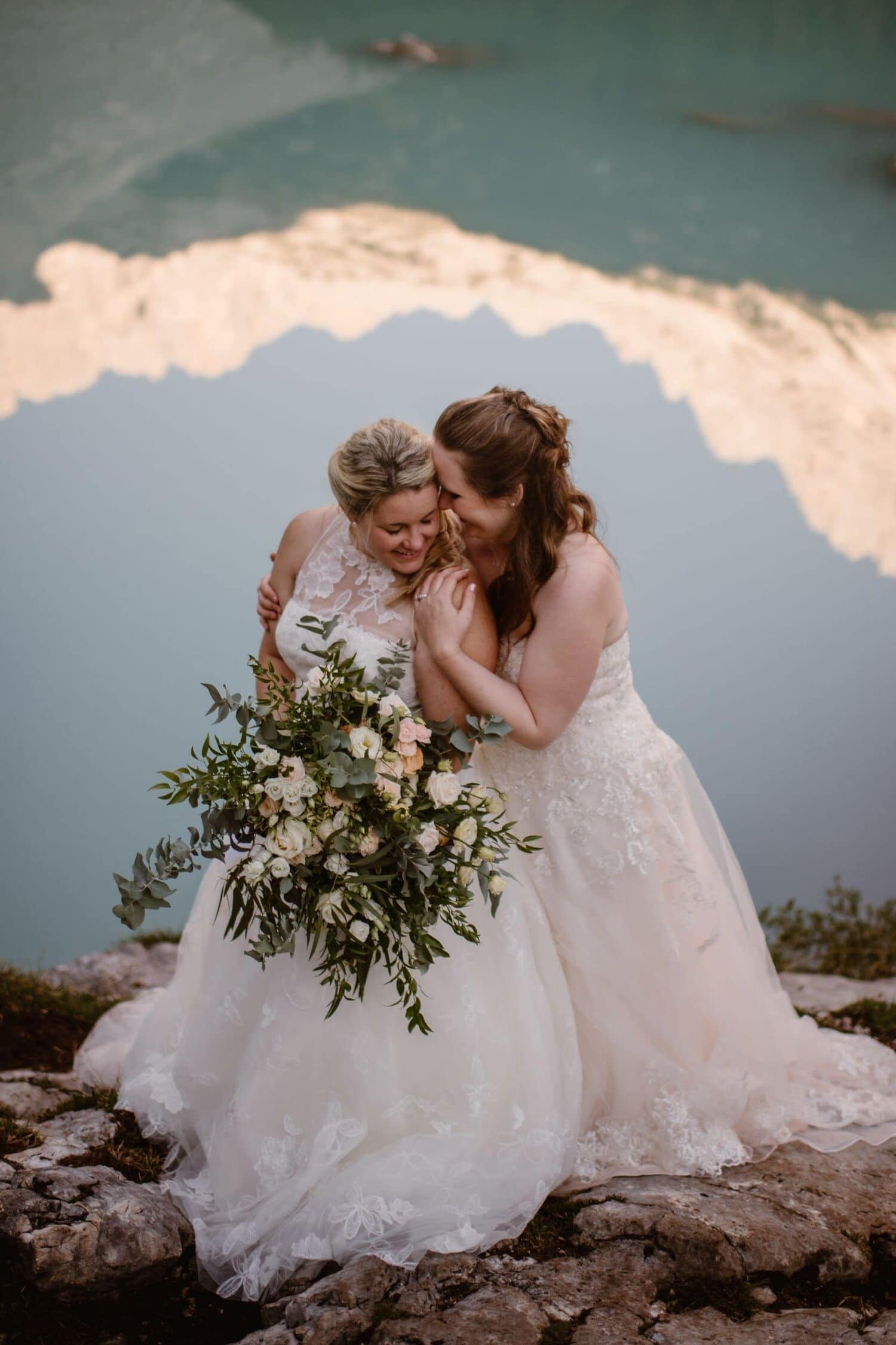 Two brides embrace and smile at each other in front of a lake.