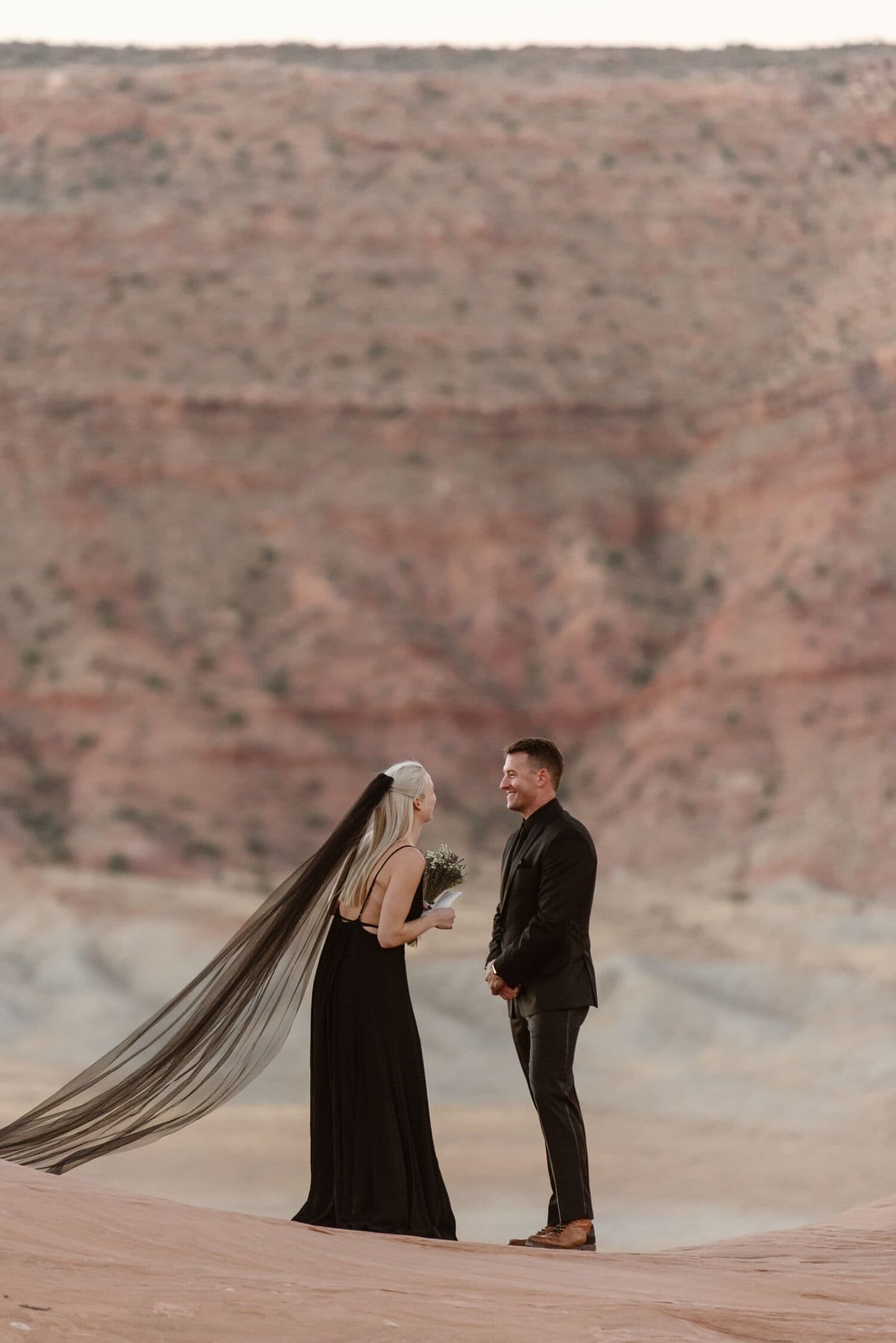 Bride and groom smile at each other during their wedding ceremony in Moab, Utah. They are wearing black wedding attire.