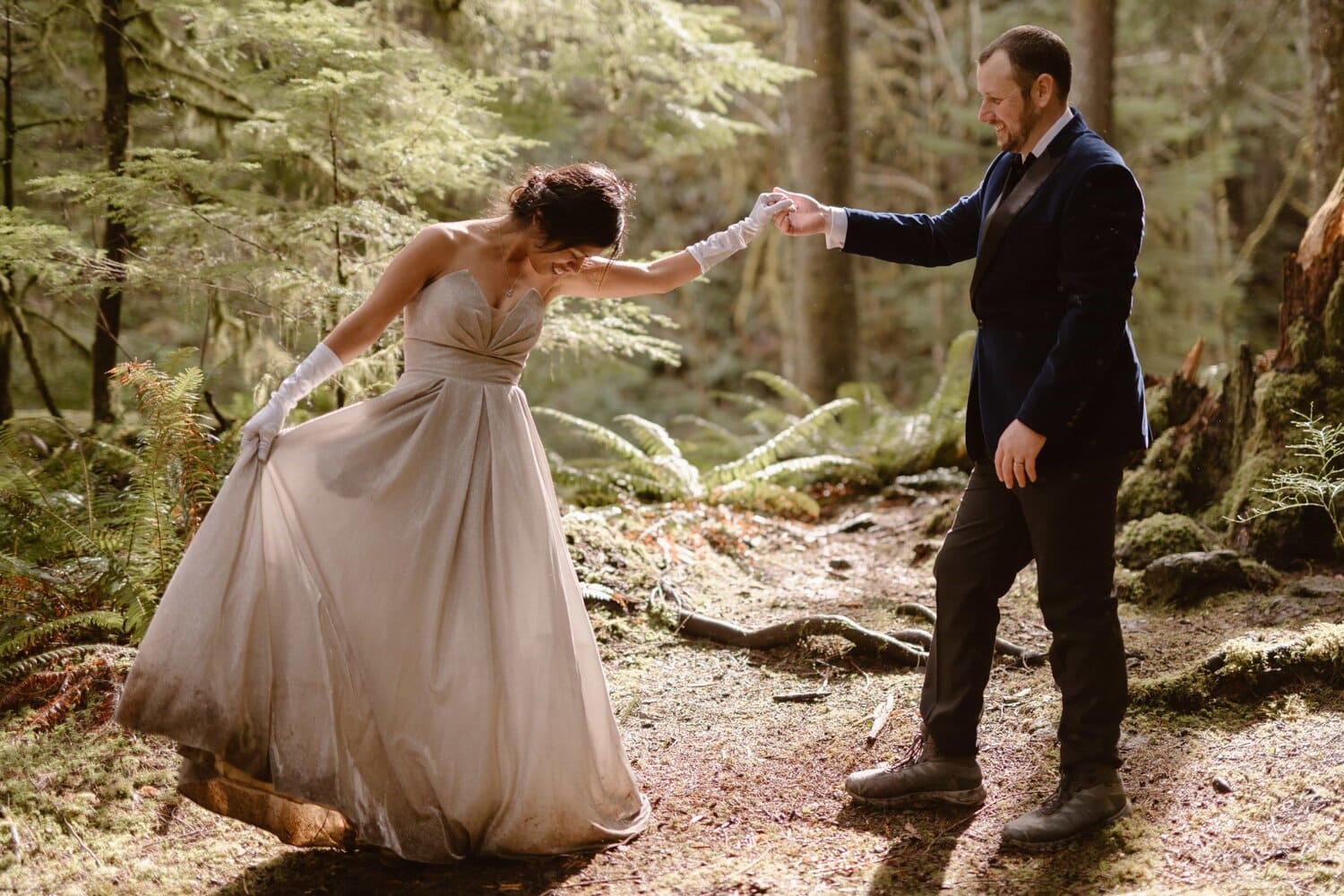 A groom holds his bride's hand in a forest. They both look at the bride's dress.