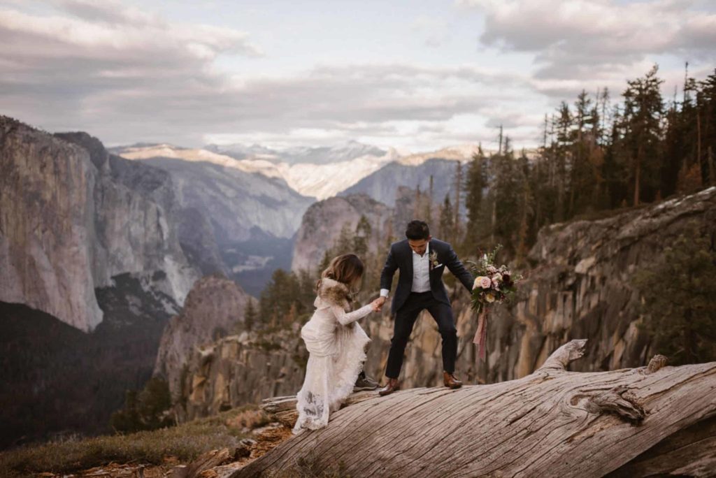 Bride and groom climb onto a fallen tree, with mountains in the background, in Yosemite National Park. 