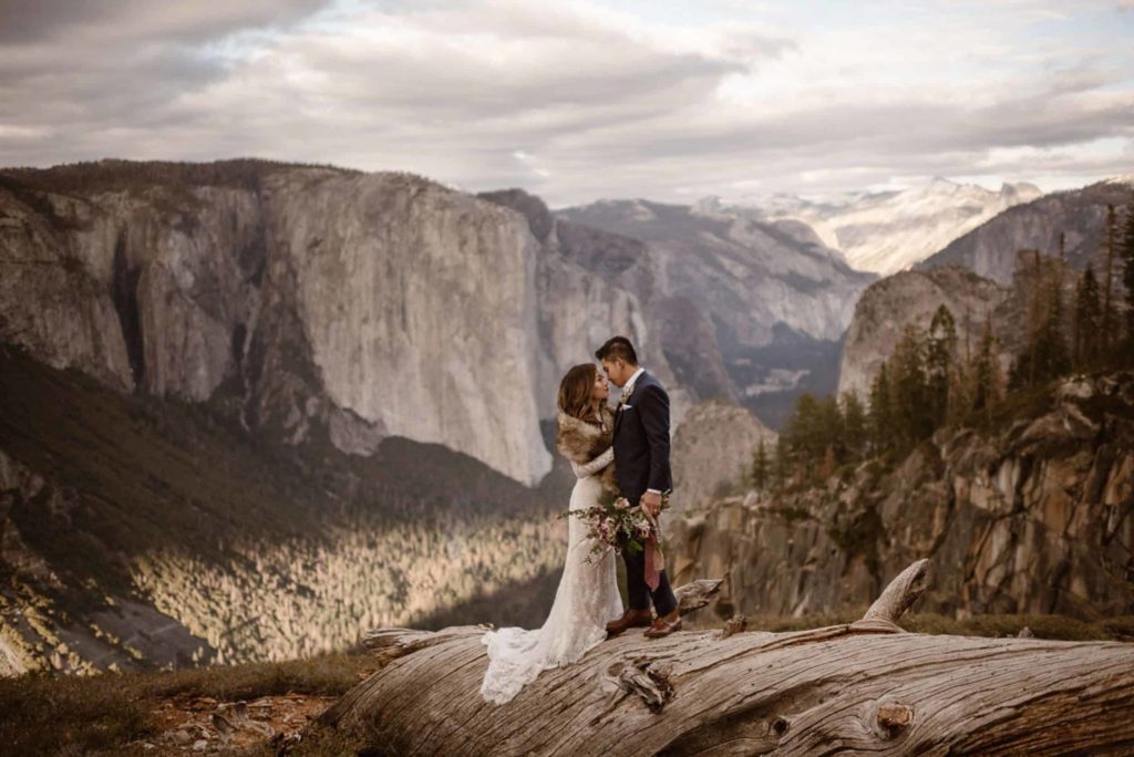 Bride and groom standing on a fallen tree, with mountains in background, in Yosemite National Park.