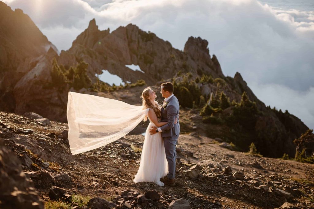 Bride and groom embrace as her white cape flows behind her. There are mountains and fog in the background. 