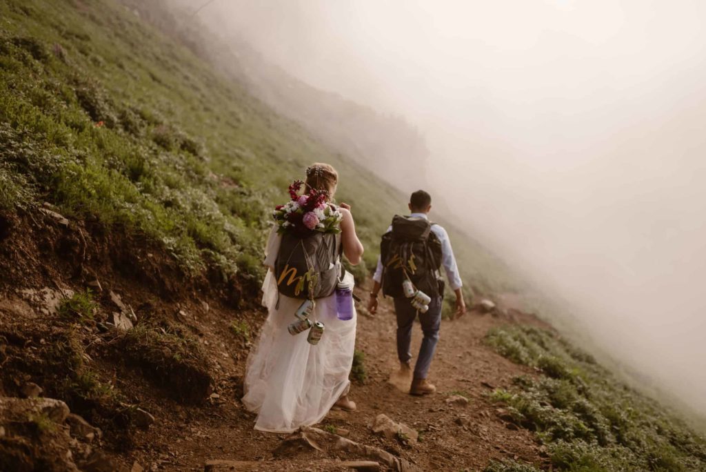 Bride and groom hiking down trail into a heavy fog, with beer cans hanging from their backpacks. 