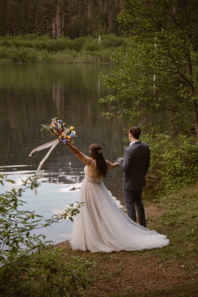 Bride and groom look out at lake, forest, and mountains in Washington State Forest. Bride holds up her bouquet.