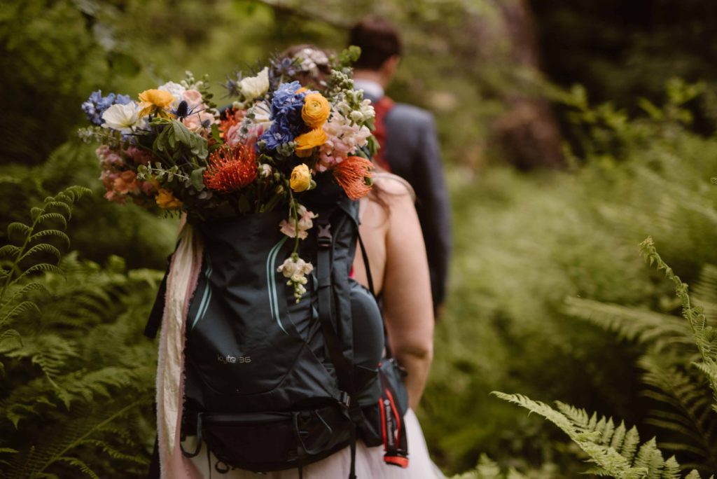 Bride and groom hike on trail surrounded by green ferns. Bride's bouquet is tucked into her backpack. 