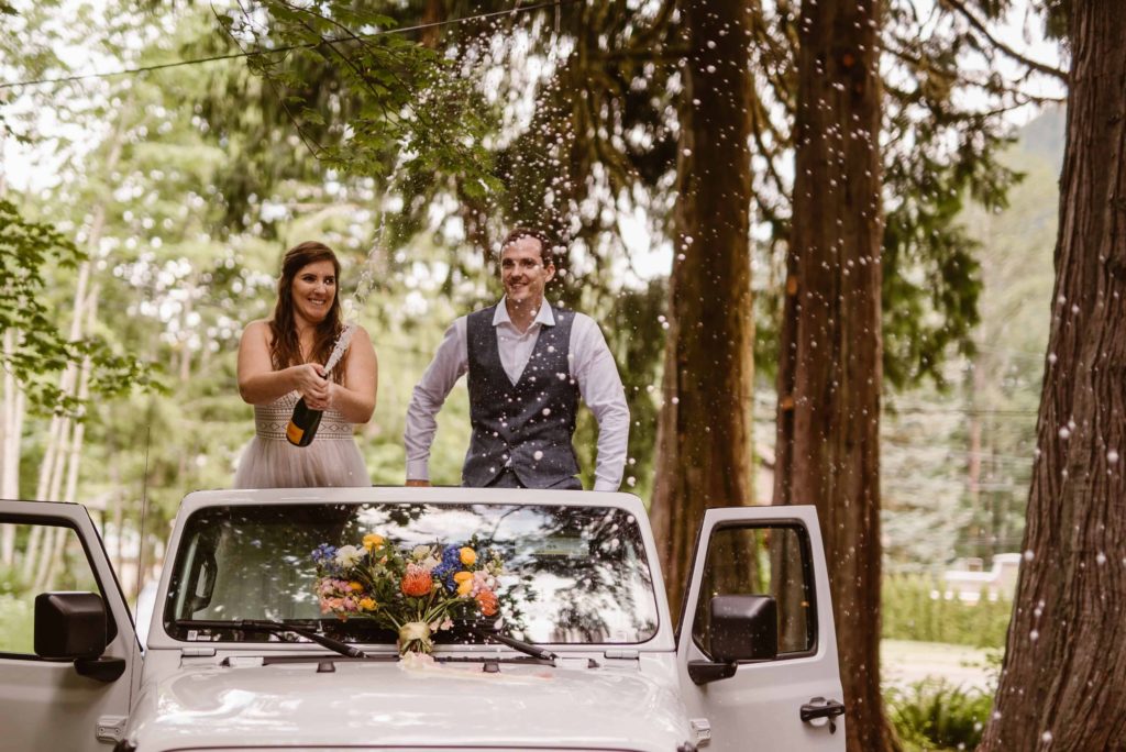Bride and groom stand in their Jeep and pop a bottle of champagne