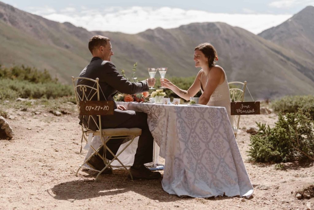 Bride and groom toast their drinks while sitting at a decorated table outside, with mountains in the background. 