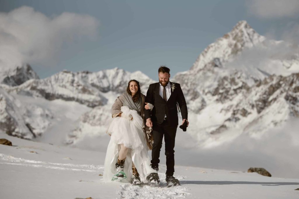 Bride and groom snowshoeing, with snow-covered mountains behind them. 
