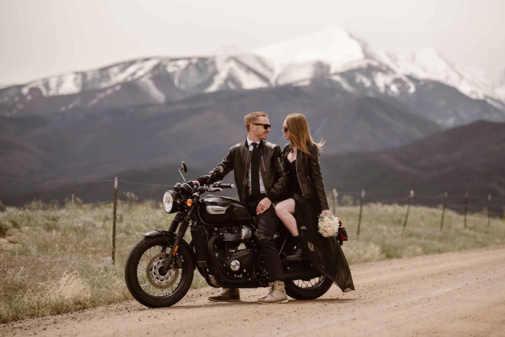 Bride and groom sitting on a black motorcycle, both wearing black leather jackets and sunglasses, with snow-capped mountains behind them. 