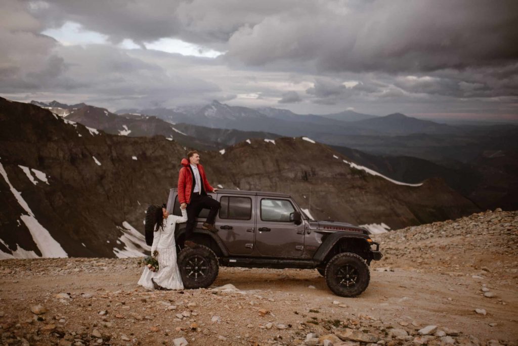 Groom stands on Jeep, while bride holds his hand below him. There are mountains in the background. 